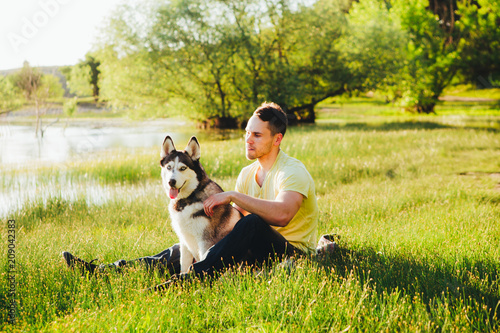 A young man is sitting on the grass with a hussy breed dog.