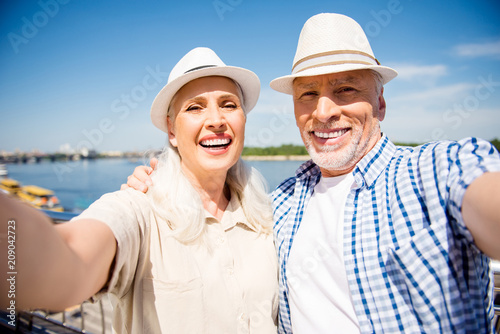 Portrait of stylish trendy senior couple in casual outfits straw hats shooting selfie on front camera over blurred river background having good mood enjoying sunny day sunshine