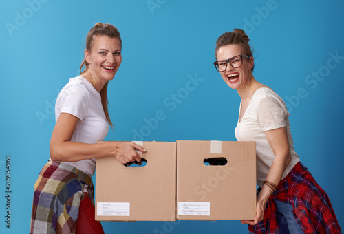 smiling modern female roommates with cardboard boxes on blue