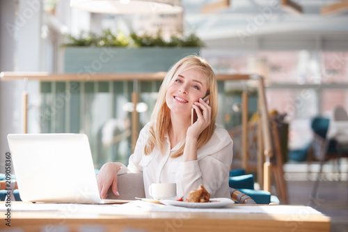 Happy young businesswoman or manager of large company talking on the phone with friends or colleagues discussing plans for the weekend sitting at table in cafe with laptop.