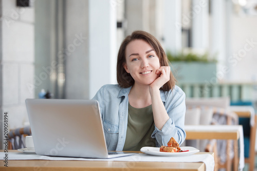 Portrait of a young charming woman looking at the camera - student or manager working with laptop at a table in an outdoor cafe.