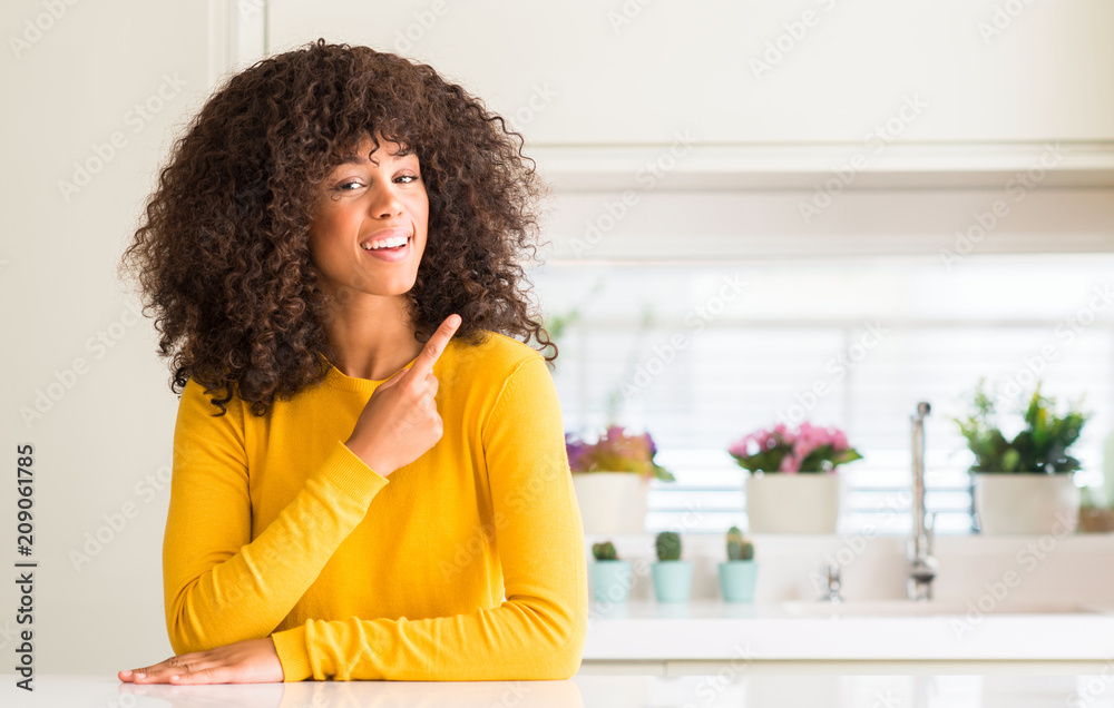 African american woman wearing yellow sweater at kitchen cheerful with a smile of face pointing with hand and finger up to the side with happy and natural expression on face looking at the camera.