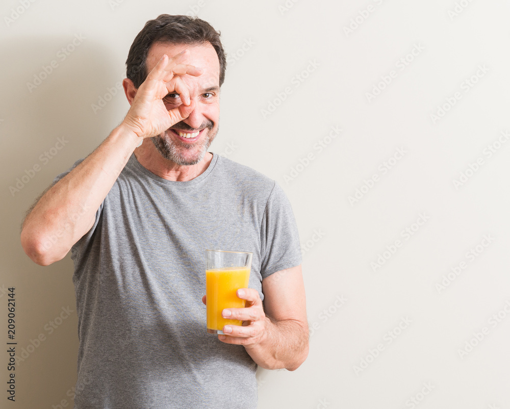 Senior man drinking orange juice in a glass with happy face smiling doing ok sign with hand on eye looking through fingers