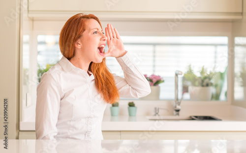 Redhead woman at kitchen shouting and screaming loud to side with hand on mouth. Communication concept.