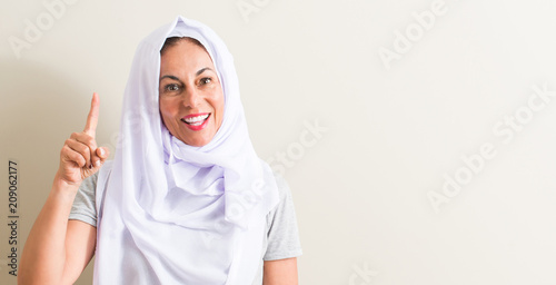 Middle age arabian woman wearing white hijab surprised with an idea or question pointing finger with happy face, number one