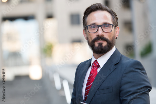 Portrait of bearded man in suit on background of building. Handsome businessman outdoor. Male business person stands on street and looking away. Businessman dressed in formal clothes and glasses..