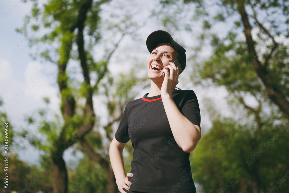 Young athletic laughing beautiful brunette girl in black uniform and cap talking on mobile phone during training, looking up and standing in city park outdoors. Fitness, healthy lifestyle concept. Young athletic laughing beautiful brunette girl in black uniform and cap talking on mobile phone during training, looking up and standing in city park outdoors. Fitness, healthy lifestyle concept.