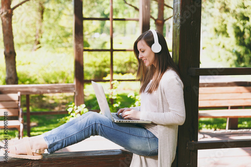 Free Download Young businesswoman in light casual clothes. Woman working on modern laptop pc computer, listen music in headphones on head in street outdoors. Mobile Office. Freelance business concept. Side view. - GFXLoad