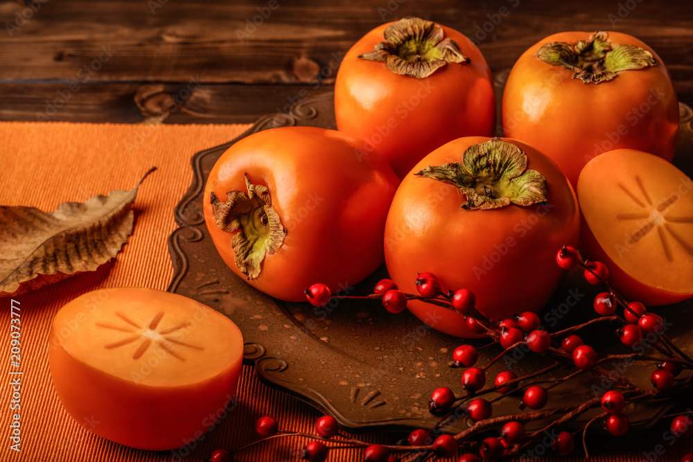 Still life of whole and halved persimmons, in a brown plate, on a wooden background.