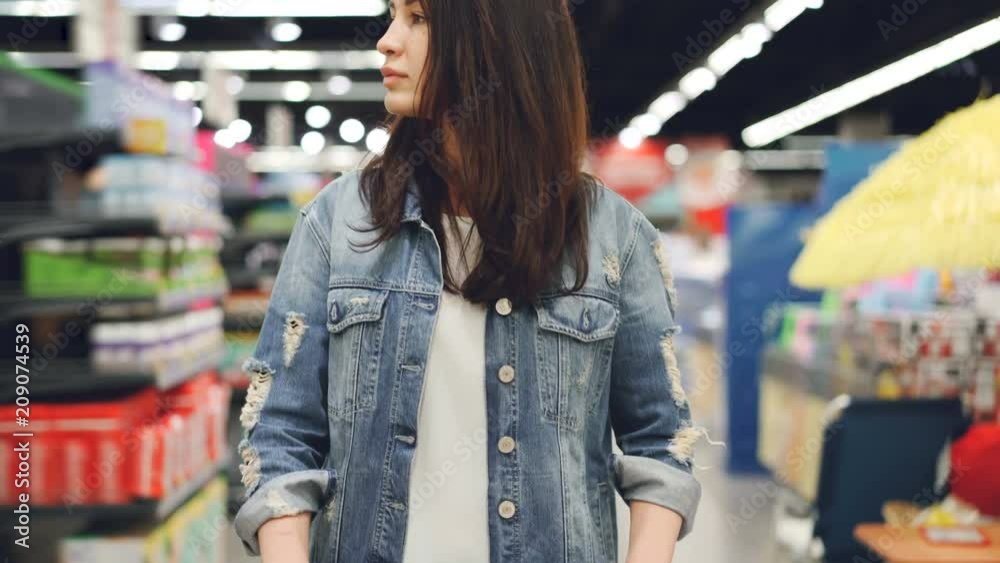 Attractive lady is going through aisle in hypermarket with shopping cart looking at shelves with bright goods. Shopping alone, beautiful people and consumerism concept.