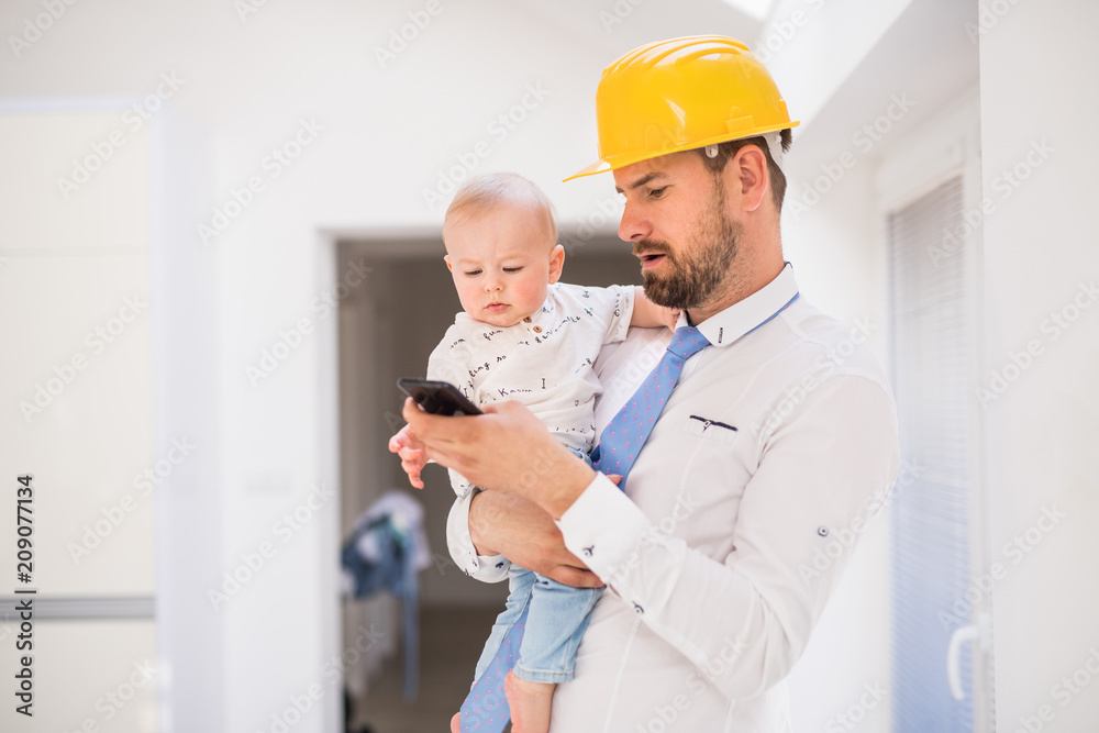 Father with shirt, tie, helmet and smartphone holding a baby son at home.