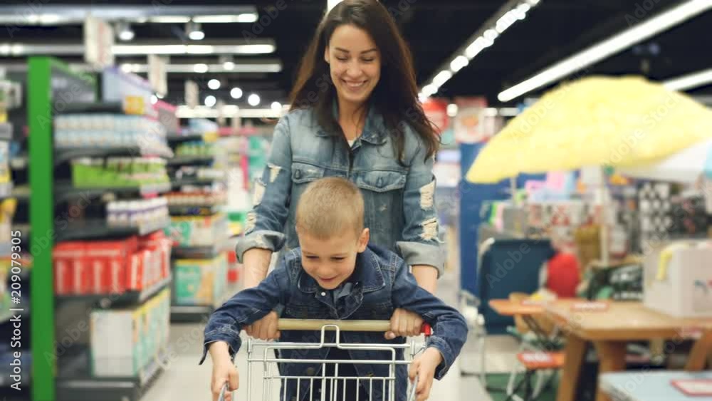 Joyful loving mother is having fun in supermarket with her cute little son, she is running with shopping cart with small boy standing on it, people are laughing.