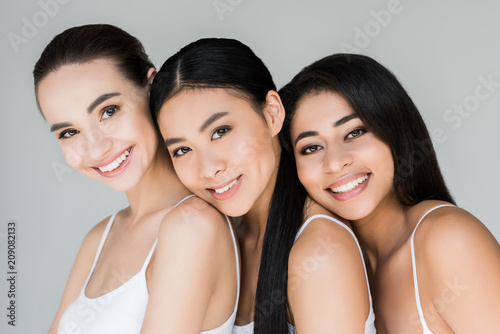 attractive smiling multiethnic women looking at camera isolated on gray background