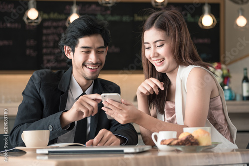 Cute couple enjoying watching on mobile phone together in cafe with coffee  bread  laptop on the table.