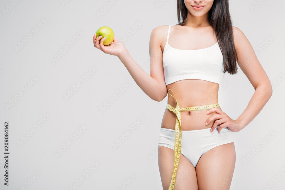 cropped view of young slim woman in lingerie with measuring tape on waist holding apple, isolated on grey, diet concept cropped view of young slim woman in lingerie with measuring tape on waist holding apple, isolated on grey, diet concept