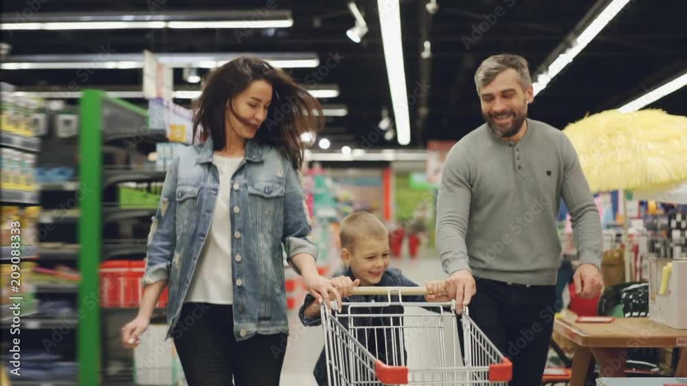 Slow motion of happy family father, mother and child running through supermarket with shopping cart, smiling and laughing. Having fun in shop and people concept.