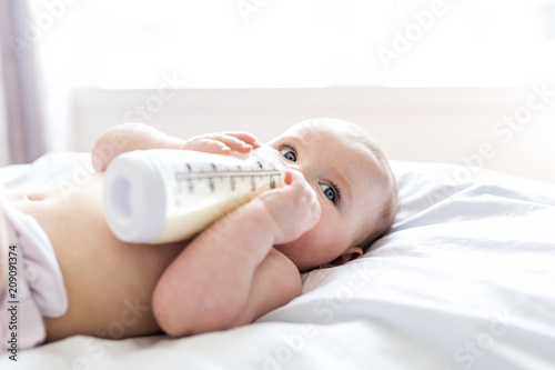 Pretty baby girl drinks water from bottle lying on bed. Child weared diaper in nursery room.