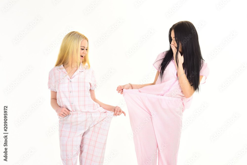 Girls on happy face shows big size of her pants. Lose weight together concept. Women, attractive young sisters, friends in pajamas. Girls on diet lost lot of weight, isolated on white background Girls on happy face shows big size of her pants. Lose weight together concept. Women, attractive young sisters, friends in pajamas. Girls on diet lost lot of weight, isolated on white background