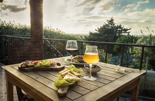 Appetizer for two on a terrace at sunset. Landscape view.  Beer glass, wine glass, cheese, olives, bread, lettuce and ham on a wooden table