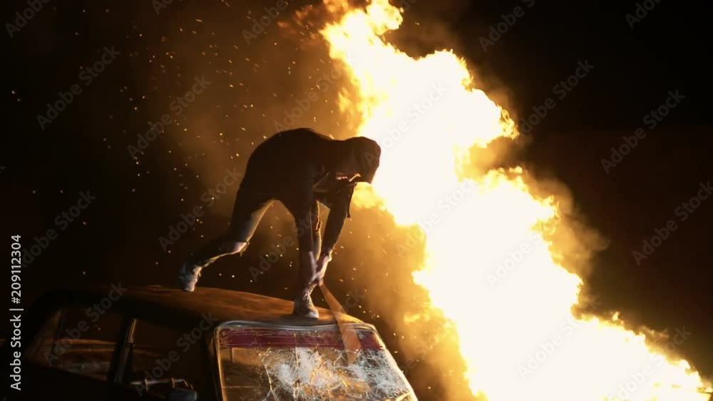 Revolution destructive behavior guy on the car with a baseball bat breaks glass. In the background is a fire. shooting at night