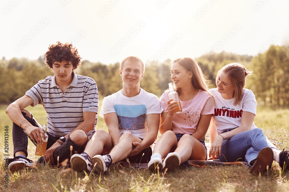 Fototapeta premium Four positive friends have joy together, have happy expressions, sit on green grass outdoor, enjoy spare time during summer vacation, pose outdoor at nature. People, friendship and recreation concept