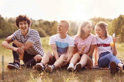 Teenagers, lifestyle and recreation concept. Four cheerful female and male companios have pleasant talk with each other, drink beer or cider, have happy expressions, share latest news, enjoy picnic © sementsova321