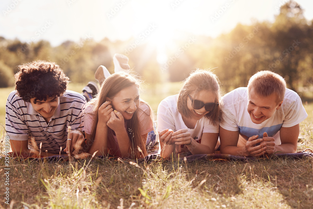 People, lifestyle, recreation and rest concept. Four joyful friends lie on  ground, have happy expressions, tell funny life stories enjoy good rest  keep legs raised. Positive two girls and boys outdoor Stock