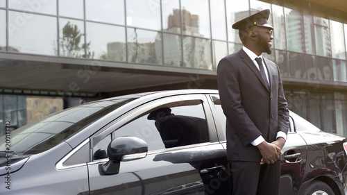 Cheerful Afro-American taxi service driver expecting passengers near luxury car © motortion
