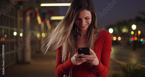 Millennial woman texting on smartphone looking happy outside in street in the evening, Caucasian girl in her 20s happily texting on phone standing on sidewalk at night, 4k