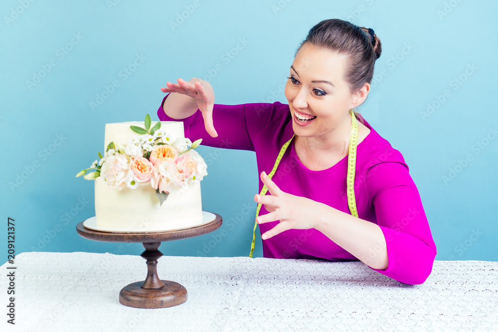 hungry and funny housewife confectioner woman looks with an appetite for a high-calorie wedding cake with measuring tape around the neck in studio on a blue background. concept of diet
