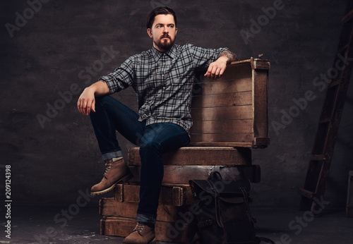 Full body portrait of a tattoed middle age hipster man dressed in a checkered shirt and jeans, sitting on wooden boxes in a studio.