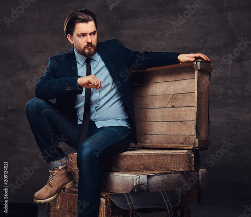 Portrait of a middle age businessman with stylish hair and beard dressed in jeans, jacket and tie, sitting on wooden boxes in a studio.