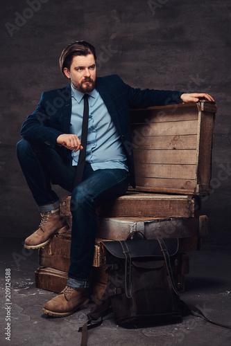 Portrait of a middle age businessman with stylish hair and beard dressed in jeans, jacket and tie, sitting on wooden boxes in a studio.