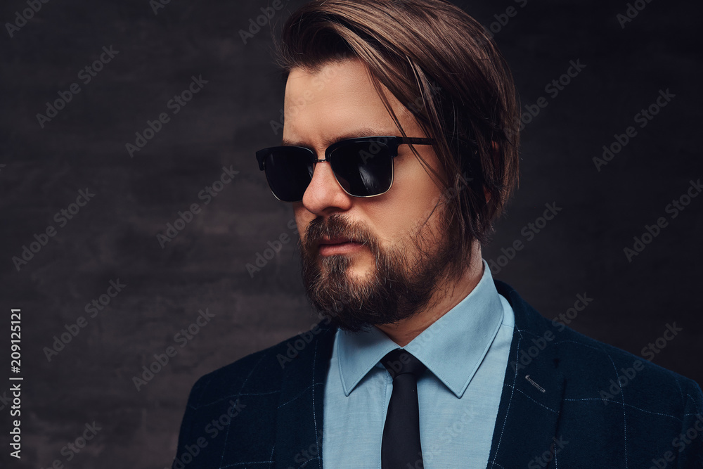 Close-up portrait of a handsome fashionable middle-aged man with beard and hairstyle dressed in an elegant formal suit and sunglasses. Isolated on a textured dark background in studio. Close-up portrait of a handsome fashionable middle-aged man with beard and hairstyle dressed in an elegant formal suit and sunglasses. Isolated on a textured dark background in studio.