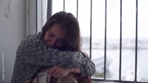 Young depressed woman is shouting at the smart phone and crying sitting on the floor by the window with rain falling outside. Lonely woman in despair with the suicide thoughts.