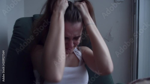 Slow motion of young depressed woman pulling her hair with hands and crying sitting in chair by the window. Raining outside. Lonely woman in despair with suicide thoughts.