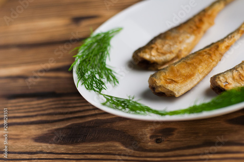 Row of three fried tasty fishes and twigs of fresh green dill on white ceramic dish on old wooden rustic brown table