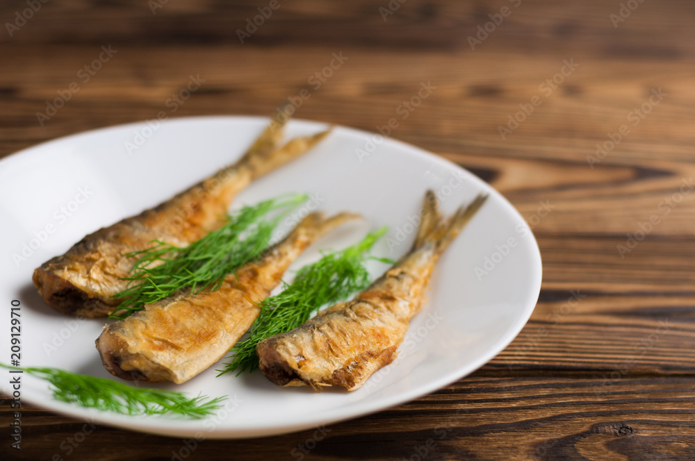 Row of three fried tasty fishes and twigs of fresh green dill on white ceramic dish on old wooden rustic brown table with copy space for your text Row of three fried tasty fishes and twigs of fresh green dill on white ceramic dish on old wooden rustic brown table with copy space for your text