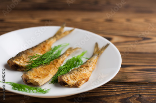 Row of three fried tasty fishes and twigs of fresh green dill on white ceramic dish on old wooden rustic brown table with copy space for your text