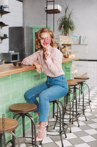 Young pretty lady in shirt and jeans sitting at the bar counter and covering her eye with lollipop candy while joyfully looking in camera in cafe