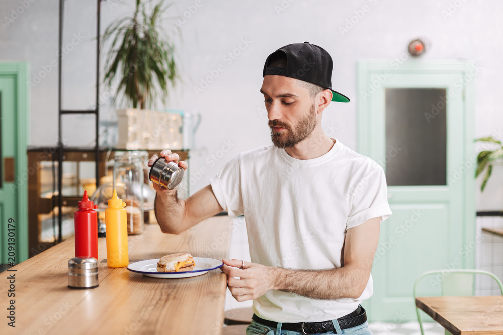 Young thoughtful man in black cap sitting at the bar counter and holding salt while eating burger in cafe Young thoughtful man in black cap sitting at the bar counter and holding salt while eating burger in cafe