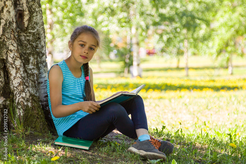 Cute little girl sitting on a green grass in summer city park and holding open book in her hands © zabavna