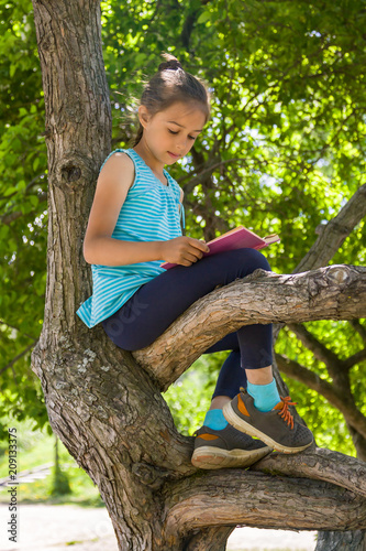 Cute little girl is sitting on a tree in summer park and reading a book 