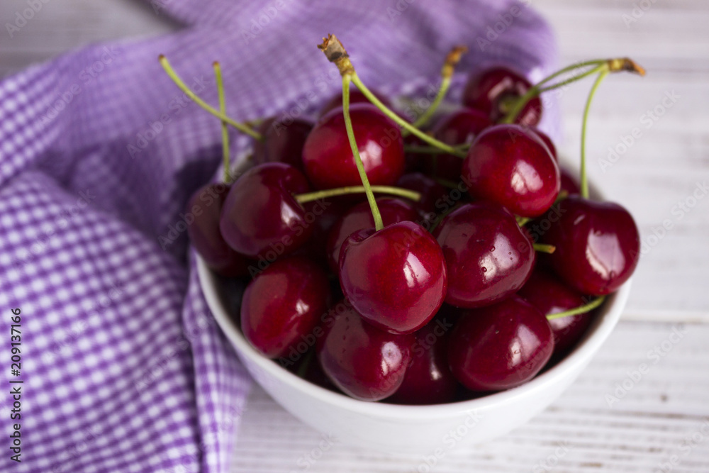 Ripe cherry berries in a white plate on a white wooden background, lilac in a cage towel. Vegetarian concept, food. Ripe cherry berries in a white plate on a white wooden background, lilac in a cage towel. Vegetarian concept, food.