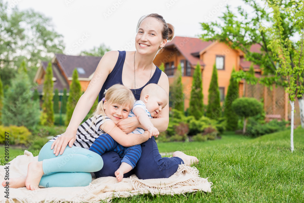 Mom with children sitting on a green grass lawn in park. Young mother with daughter and son having fun on picnic at backyard on bright sunny day. Happy family enjoy spending time outdoors concept Mom with children sitting on a green grass lawn in park. Young mother with daughter and son having fun on picnic at backyard on bright sunny day. Happy family enjoy spending time outdoors concept