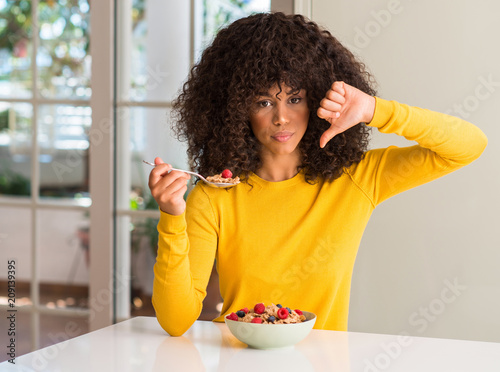 African american woman eating cereals, raspberries and blueberries with angry face, negative sign showing dislike with thumbs down, rejection concept