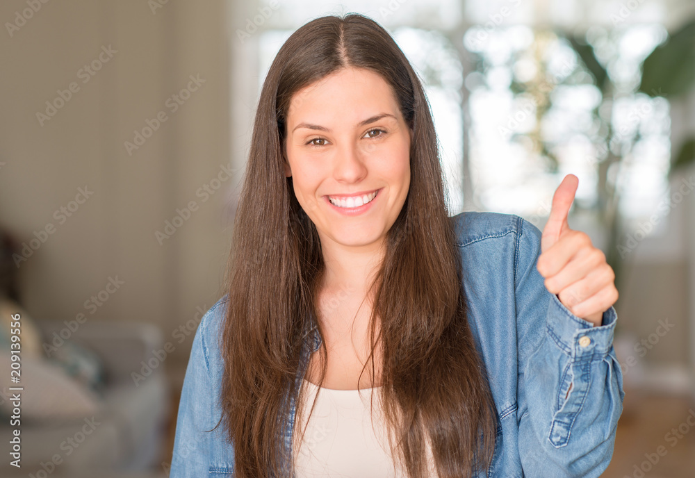 Young beautiful woman at home happy with big smile doing ok sign, thumb up with fingers, excellent sign Young beautiful woman at home happy with big smile doing ok sign, thumb up with fingers, excellent sign