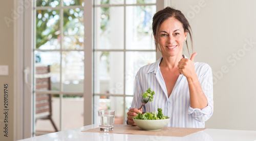 Middle aged woman eating fresh salad in a bowl at home happy with big smile doing ok sign, thumb up with fingers, excellent sign