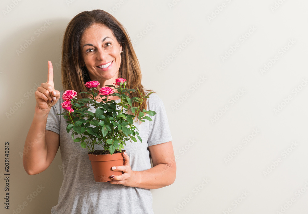 Middle age woman holding roses flowers on pot surprised with an idea or question pointing finger with happy face, number one Middle age woman holding roses flowers on pot surprised with an idea or question pointing finger with happy face, number one