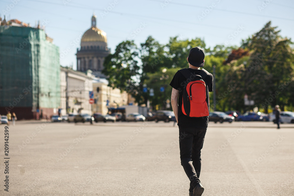 Back view of young stylish man tourist walking at Palace square near Saint Isaac's Cathedral in Saint-Petersburg. He wearing in black jeans, black T-shirt, black cap and red backpack